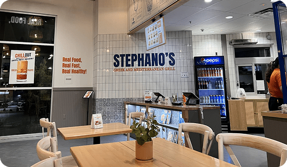 Locations 5 A restaurant interior with "STEPHANO'S GREEK AND MEDITERRANEAN GRILL" on a tiled wall and wooden tables.