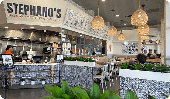 Locations 7 A brightly lit restaurant with "STEPHANO'S GREEK AND MEDITERRANEAN GRILL" above the counter and customers dining.