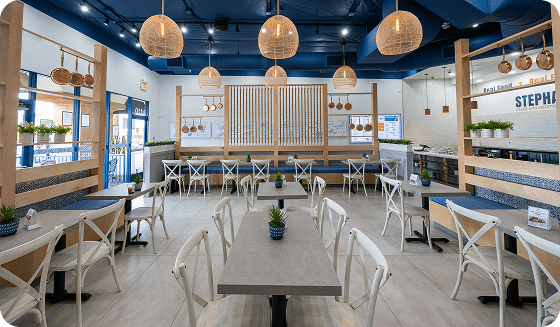 Locations 4 A modern restaurant with a blue ceiling, light wood accents, white cross-back chairs, and gray tables.