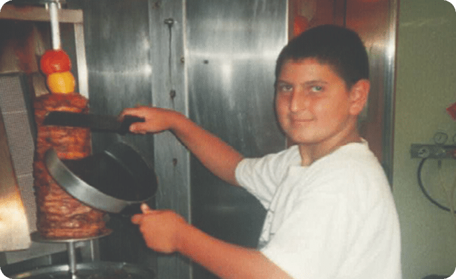 A young boy in a white t-shirt smiles while slicing meat from a vertical rotisserie onto a pan.