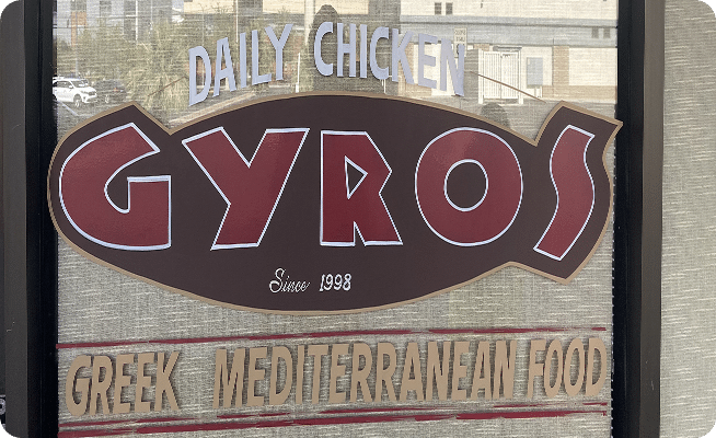 A store sign with "DAILY CHICKEN GYROS Since 1998 GREEK MEDITERRANEAN FOOD" in white, red, and brown text.