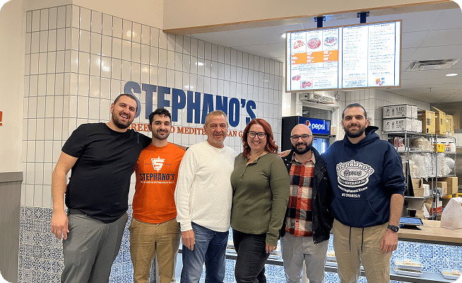 Six smiling people stand in front of a white tiled wall with "STEPHANO'S" in blue letters.