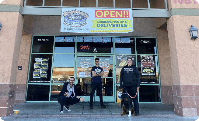 Three people and a Doberman dog pose in front of a storefront with a banner reading "OPEN!! Curbside Pick Up & Deliveries."