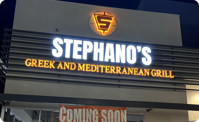 A brightly lit sign for "STEPHANO'S GREEK AND MEDITERRANEAN GRILL" with a "COMING SOON" banner below.